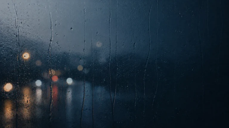 雨水沿深色窗玻璃流下，窗外街灯化成朦胧光点